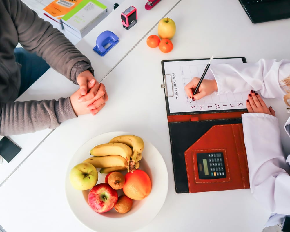Consulta de nutrición presencial: plan de alimentación semanal sobre el escritorio, junto a fruta fresca